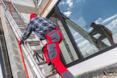 Bir inşaat işçisi, bulutlu açık mavi gökyüzünün altındaki bir inşaat sahasındaki görevine odaklanmış bir şekilde merdivene tırmanır..