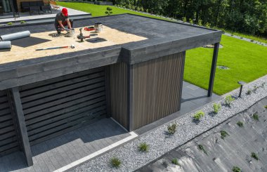 Becerikli işçiler çağdaş bir binanın çatısında bakım işlerini tamamlıyorlar. Yakınlarda yemyeşil bir yer ve etrafa saçılmış aletler var..