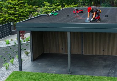 Bir inşaat işçisi düz çatılı bir yapıya çatı malzemesi uyguluyor. Çevresi yeşillik ve aletlerle çevrili. Arka bahçede..