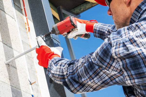 A construction worker in a plaid shirt and gloves attaches metal framing to a building's exterior with a power drill under clear blue skies.