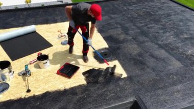 İşçi güneşli bir günde çatıya dolgu macunu sürüyor. Bakım ve onarım için çatıya çeşitli araç ve gereçler yerleştiriliyor..