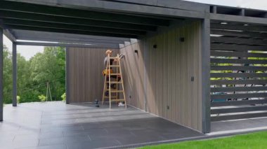 Bir işçi, çevresi yeşilliklerle çevrili çağdaş bir garaj binasına ahşap paneller çizmek için merdivene tırmanır. Ortalık sakin ve huzurlu..