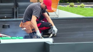 Yetenekli bir işçi arka bahçeye yeni bir çatı kurarken EPDM çatı malzemesine ısı uygular. Çevrede modern açık hava mobilyaları ve yeşillikler yer almaktadır..