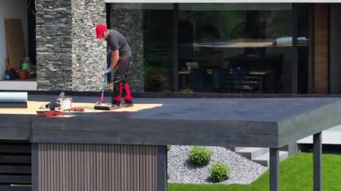Bir işçi banliyö bölgesindeki bir evin çağdaş düz çatısına EPDM çatı malzemesi uyguluyor, gün ışığında inşaat becerilerini sergiliyor.