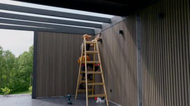 Yetenekli bir işçi, öğleden sonra merdiveni kullanarak çağdaş bir ahşap yapının dış yüzeyine son rötuşları dikkatlice uygularken görülüyor..