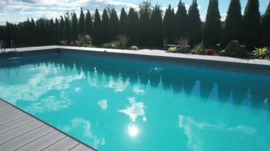 Sakin bir yüzme havuzu, parlak bir gökyüzünün altında, yemyeşil ve bahçelerle çevrili, berrak mavi bir su sergiliyor..