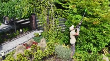 Bir bahçıvan, temiz gökyüzünün ve parlak güneş ışığının tadını çıkarırken iyi korunan bir yerleşim alanındaki çalıları ve ağaçları buduyor..