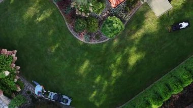 İki profesyonel, temiz bir gökyüzünün altında ağaçlar ve bitkilerle dolu güzel bir banliyö bahçesinde çimleri ve çitleri kesiyor..