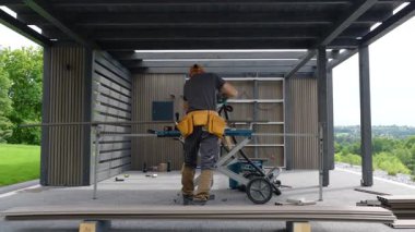 Yetenekli bir marangoz, arka planda yeşillik olan çağdaş bir açık alanda çerçeve inşa ederken malzemeyi kesmek için testere kullanır..