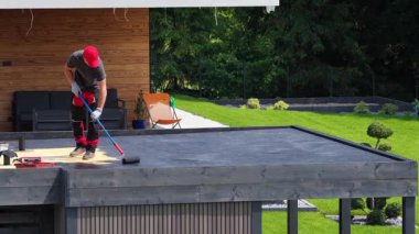 Becerikli bir işçi, güneşli bir banliyö ortamında düz bir çatıya koruyucu bir kaplama uyguluyor, uzman tekniği ve kullanılan aletleri sergiliyor..