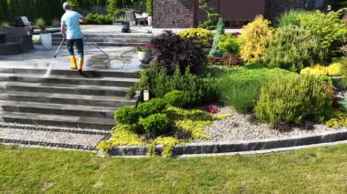 Parlak giysili şahıs bahçe merdivenlerini temizler. Etrafı yemyeşil ve peyzajla çevrilidir. Açık havada güneşli bir gün geçirir..