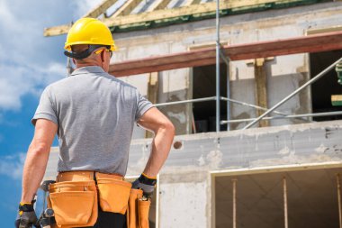 Sarı başlıklı inşaat işçisi inşaat alanını inceliyor. İnşaat iskelesi ve tamamlanmamış duvarlar görünür, gündüz devam eden çalışmalar.