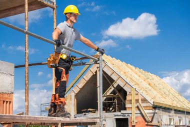 Bir inşaat işçisi, ahşap bir yapının altında kabarık bulutlu parlak mavi bir gökyüzü inşa edilirken, iskelenin üzerinde durup, alanı gözlemliyor..