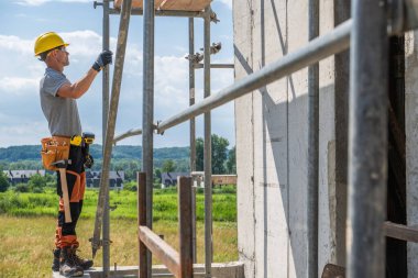 İnşaat işçisi inşaat iskelesini inceliyor, yeşil tarlalar ve mavi gökyüzü ile çevrili. Çalışma ortamında güvenlik tedbirleri belirgindir.