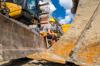 Kasklı bir inşaat işçisi, bulutlu açık mavi bir gökyüzünün altında yoğun bir inşaat sahasında ağır makineler üzerinde otururken mola verir..