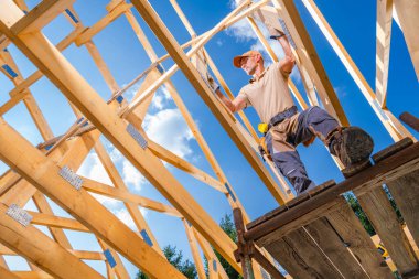 Bir marangoz ahşap kirişleri bir çatı için birleştirirken ustalıkla dengede durur parlak mavi gökyüzü altında, kabarık bulutlar, gösterişli zanaatkarlık.