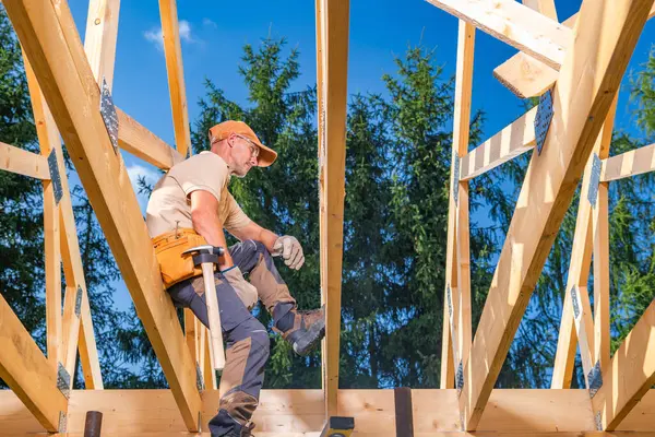 Bir marangoz açık mavi gökyüzünün altındaki ağaçların arasında ahşap çerçeveler üzerinde denge kurarken aktif olarak çatı destek kirişleri inşa eder. Güvenlik ekipmanları kullanımda.