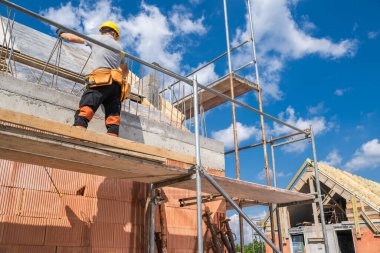 Bir inşaat işçisi yeni bir bina için tuğlalar döşeyerek, yapı iskelesi ve açık mavi gökyüzü ile çevrili olarak gayretle çalışıyor..