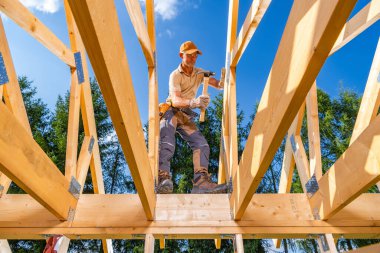 Yetenekli bir işçi, gün ışığında, yüksek ağaçlar ve açık mavi gökyüzü ile çevrili bir inşaat alanında ahşap çerçeveler kurmaya odaklanır..