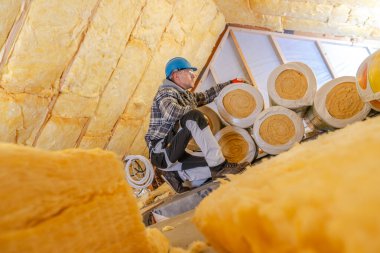 Miğfer ve ekose gömlekli bir işçi, gündüz yenileme projesi sırasında güvenlik ve hassasiyete odaklanan bir çatı katına izolasyon yerleştirir..