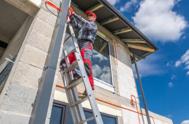 Kareli gömlekli ve kırmızı pantolonlu bir inşaat işçisi mavi gökyüzünün altında kabarık bulutlu bir binaya malzeme yüklemek için merdivene tırmanır..
