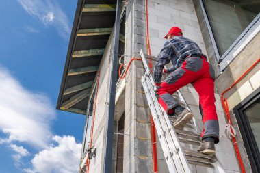 Kırmızı pantolonlu bir işçi gün ışığında bir binayı incelemek için merdivene tırmanır. İnşaat alanında açık duvarlar ve açık gökyüzü yer alıyor.
