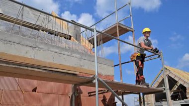 Bir inşaat işçisi, açık mavi gökyüzünün altında ahşap iskeleti olan bir bina projesini denetleyerek, bir yapı iskelesinde güven içinde duruyor..