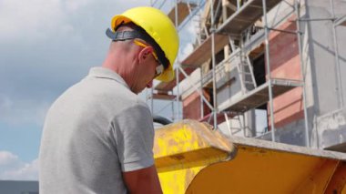 Sert şapka takan bir inşaat işçisi, açık mavi gökyüzünün altındaki hareketli bir inşaat alanında proje için malzemeleri karıştırıyor..