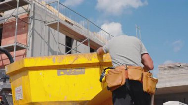 Gri gömlekli ve alet kemerli bir inşaat işçisi açık mavi gökyüzünün altındaki bir inşaat alanındaki parlak sarı bir çöp konteynırına malzeme yerleştiriyor..