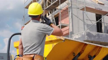 Bir inşaat işçisi açık bir gökyüzünün altındaki inşaat alanında kask ve eldiven giyerken bir inşaat projesini denetliyor..