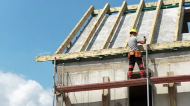 Bir işçi inşaat halindeki bir binaya yeni çatı panelleri kurmakla meşgul. Parlak gökyüzü ve yapı iskelesi dinamik bir çalışma ortamı yaratır..