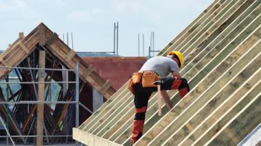 Yetenekli işçiler, güneşli bir günde bir şantiyede ahşap iskelet inşa ederken güvenlik teçhizatı kullanarak çatı kirişlerine tırmanıyor..