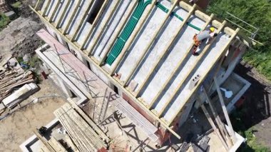 Çevredeki inşaat devam ederken, işçiler yeni bir evin çatı yapısını kuruyorlar. Araçlar ve malzemeler belirgin bir şekilde kullanımda.