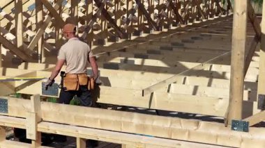 Bir inşaat işçisi bir ev ahşap iskeleti üzerinde duruyor, dikkatle ölçüyor ve yeşil alanlarla çevrili bir kırsal alanda yerleşimi denetliyor..