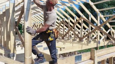 Bir marangoz, bir inşaat alanında bir binanın çatı yapısını birleştirmek için özenle çalışır. Açık mavi gökyüzü görevin enerjik atmosferine eklenir.