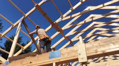 Bir marangoz, açık mavi gökyüzünün altındaki ahşap kirişlerin üzerinde durarak evin çatı iskeletini inşa ediyor. İnşaat alanında aydınlık ve yoğun bir gün..