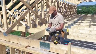 Bir inşaat işçisi, açık gökyüzünün altındaki inşaat sahasında ev kirişlerini güvence altına almak için elektrikli aletler kullanırken eldiven ve şapka takıyor..