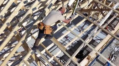 Bir inşaat işçisi açık gökyüzünün altındaki bir yerleşim alanında ustalıkla ahşap iskelet monte eder. Güvenlik teçhizatı yerin üstündeyken kullanılıyor..