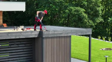 Bir işçi, yüzeyi son rötuşlara hazırlayan bir silindir kullanarak düz bir çatı güvertesine boya sürer. Yeşillik ve berrak gökyüzü canlı bir zemin yaratır..