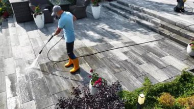 Parlak sarı çizmeli bir insan, güneşli bir öğleden sonra taze çiçekler ve yeşilliklerle çevrili bir taş verandayı temizlemek için basınçlı çamaşır makinesi kullanır..