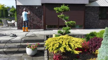 Aydınlık ve güneşli bir günde evlerin dış yüzeyini iyileştirirken, çeşitli bitkilerle birlikte iyi korunan bir bahçedeki peyzajlar üzerinde çalışan bir kişi..