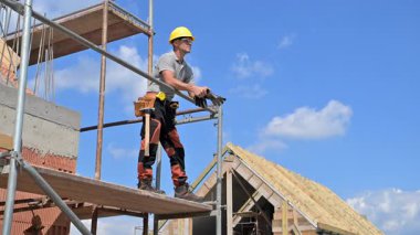 Bir inşaat işçisi yapı iskelesinde dururken malzemeleri ölçüyor. Site, üzerinde açık gökyüzü bulunan inşaat halinde yeni bir binaya sahip..