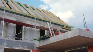 İşçiler, parçalı bulutlu bir gökyüzünün altındaki ahşap çatı iskeletine odaklanarak modern bir ev inşa etmekle meşguller. İnşaat malzemeleri ve yapı iskelesi görünür..