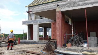 Bir inşaat işçisi, güneşli bir havada inşaat alanındaki ilerlemeyi inceliyor. Yapıda açık duvarlar ve devam eden gelişmeler yer alıyor.