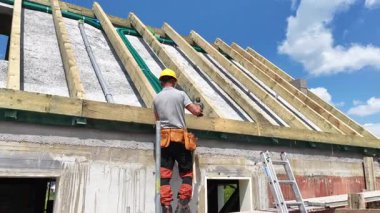 Bir inşaat işçisi yeni bir binaya çatı kirişleri yerleştirir, aletleri kullanır ve güneşli bir günde güvenlik kaskı takar..