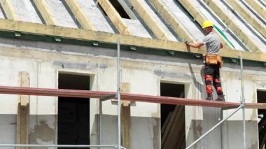 Bir inşaat işçisi, gündüz vakti yeni bir binaya ustalıkla ahşap iskelet yerleştiriyor, sıcak hava koşullarında takım çalışması ve güvenlik uygulamaları sergiliyor..