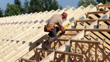 Bir inşaat işçisi çatı iskeletine ahşap kirişleri ölçer ve yerleştirir. Alan hareketli ve arka planda ağaçlar var..