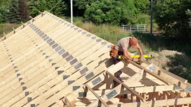 Bir inşaat işçisi, gün ışığında ağaçlarla çevrili bir eve dikkatlice çatı iskeleti kuruyor..