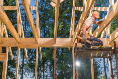 Bir marangoz bir iskele üzerinde tünemiş iken ahşap iskelet inşa etmeye odaklanır. Bölgenin etrafını saran yemyeşil ağaçlar kırsal bir bölgeyi işaret ediyor..