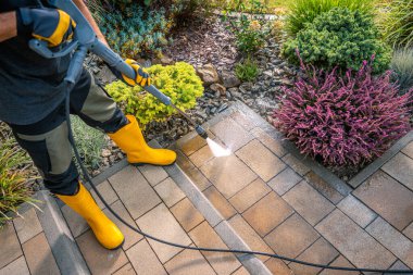 Sarı çizme giyen bir insan, parlak güneş ışığı altında canlı bitkiler ve çiçeklerle dolu iyi korunmuş bir bahçede kaldırım taşlarını temizlemek için basınç makinası kullanır..